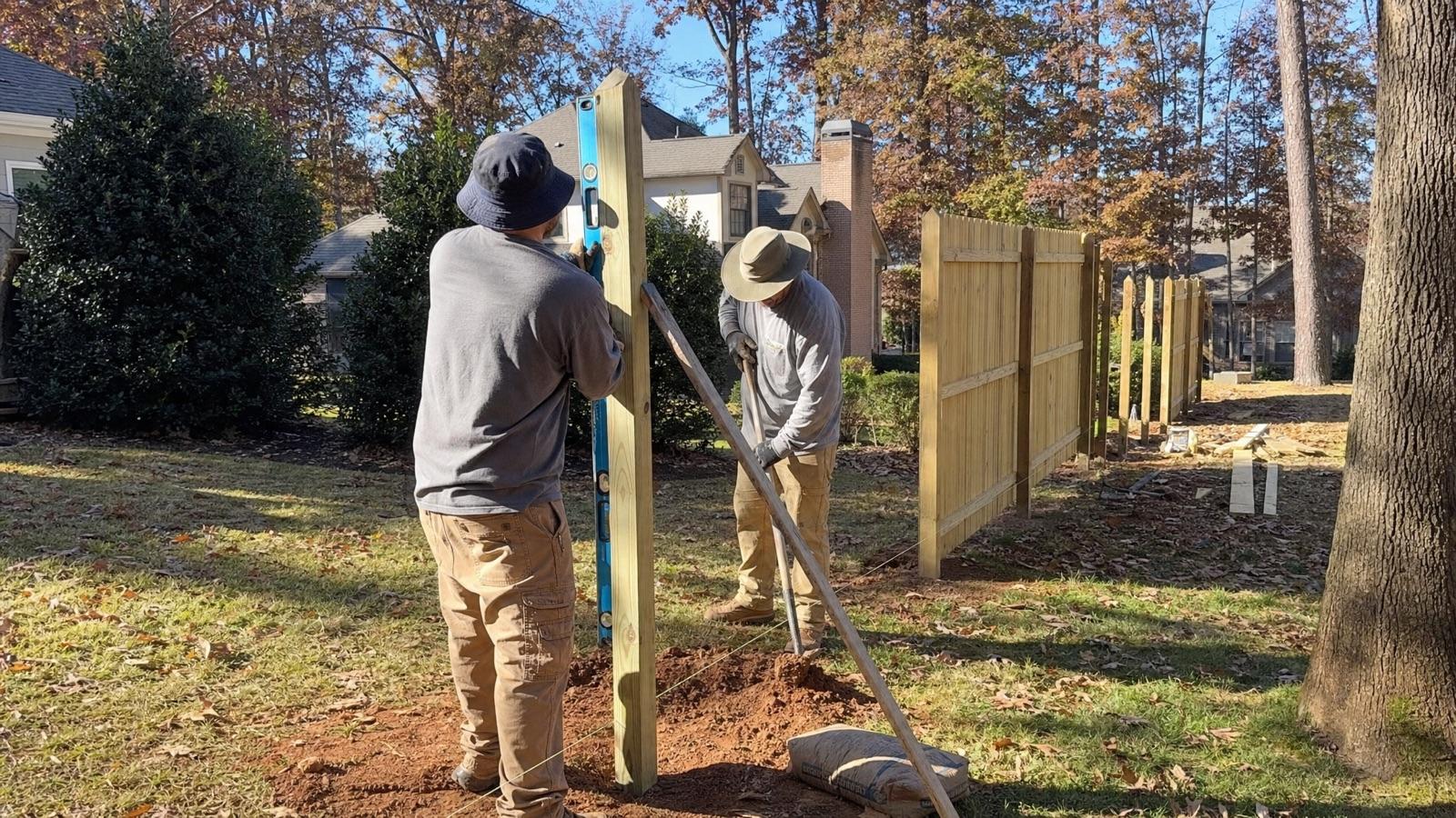 Fence installation crew setting a wood fence post in cool autumn weather along a back property line
